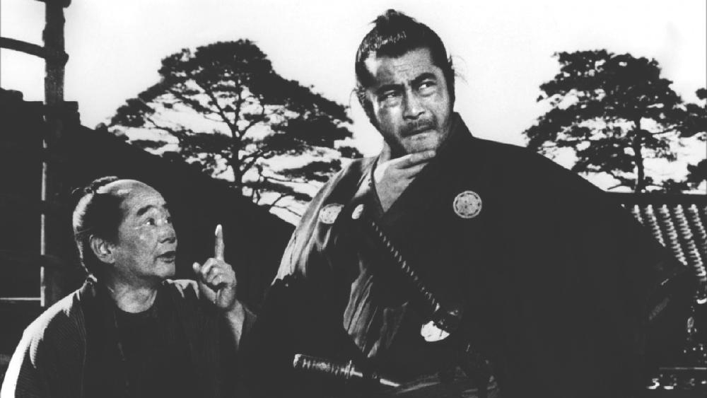 An image from the film Yojimbo of Toshiro Mifune stroking his beard, looking away, as a villager tries to speak to him.