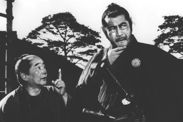 An image from the film Yojimbo of Toshiro Mifune stroking his beard, looking away, as a villager tries to speak to him.